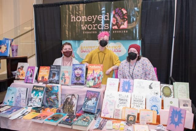 Drei Personen hinter einem Messestand mit Büchern auf einem Festival.